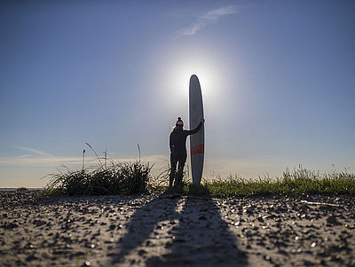 Surfer/in steht mit Surfbrett am Strand mit Blick Richtung Sonne