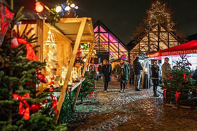 Besucher schlendern am Abend über einen festlich beleuchteten Weihnachtsmarkt mit Holzhütten, geschmückten Tannenbäumen und einem Grillstand. Im Hintergrund leuchtet ein modernes Gebäude mit Glasfassade.