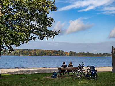Ein Mann und eine Frau sitzen auf einer Bank und blicken auf einen See. Hinter der Bank stehen zwei Fahrräder.