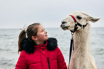 Ein Mädchen und ein Lama stehen an Meer und schauen sich an.