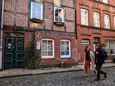 Zwei Personen spazieren durch eine historische Altstadtgasse mit Kopfsteinpflaster. Im Hintergrund prägen liebevoll erhaltene Backstein- und Fachwerkhäuser das Stadtbild.