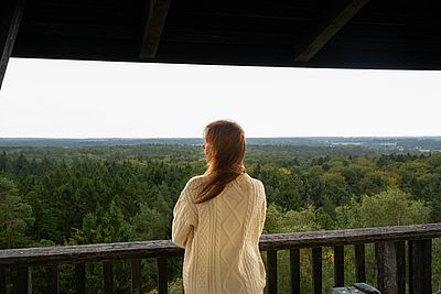 Eine Frau blickt von einem hölzernen Aussichtsturm über die weitläufige Waldlandschaft der Hahnheide.