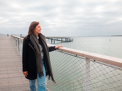 Eine Frau in dunklem Mantel und grauem Schal steht auf einer Seebrücke, hält sich am Geländer fest und blickt lächelnd über das ruhige Meer unter bewölktem Himmel.