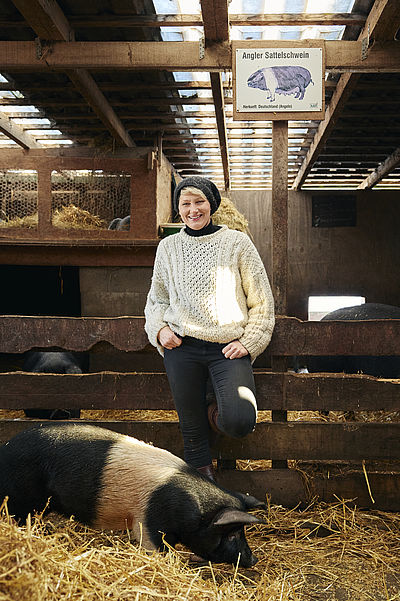  Eine Frau steht lachend auf dem Arche-Hof Bredland in einem Schweinestall, vor ihr läuft ein Schwein.