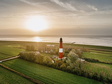 Luftaufnahme eines rot-weiß gestreiften Leuchtturms inmitten grüner Felder an der Nordseeküste. Im Hintergrund erstreckt sich das Wattenmeer, über dem die tief stehende Sonne durch die Wolken scheint.