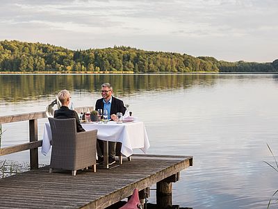Ein Mann und eine Frau sitzen an einem Esstisch auf einem Steg am See.