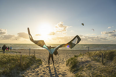 Ein Kiter steht am Strand und hält seinen Kite-Schirm im Wind, während die Sonne untergeht.