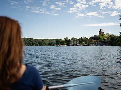 Eine Frau paddelt mit einem Kajak über den Ratzeburger See. Am Ufer sind Segelboote und der Ratzeburger Dom zu sehen.
