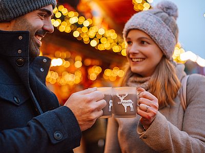 Ein lächelndes Paar in winterlicher Kleidung stößt auf einem festlich beleuchteten Weihnachtsmarkt mit zwei Tassen Heißgetränk an.