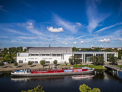 Außenansicht der Musik und Kongresshalle Lübeck. Davor sieht man den Fluss Trave und ein Theaterschiff.