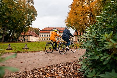 Zwei Personen fahren mit Fahrrädern auf einem Weg durch eine Parkanlage. Beide tragen Fahrradhelme und warme Jacken, eine Person eine orangefarbene, die andere eine dunkelblaue. Im Hintergrund ist ein großes Gutshaus mit rotem Dach zu sehen, umgeben von herbstlich gefärbten Bäumen.