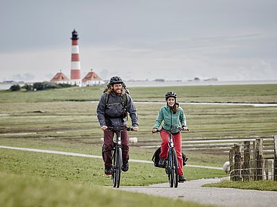 Zwei Personen fahren mit Fahrrädern durch eine weite Küstenlandschaft. Im Hintergrund erhebt sich ein rot-weiß gestreifter Leuchtturm unter einem bewölkten Himmel.