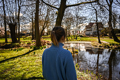 Eine junge Frau steht im Schatten eines Baumes im Gerisch Skulpturenpark und blickt auf einen Teich.