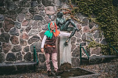 Der Till-Eulenspiegel-Darsteller Sven Kolb steht vor dem Eulenspiegel-Brunnen in Mölln. Er trägt ein Eulenspiegel-Kostüm mit Glockenmütze und Schnabelschuhen.