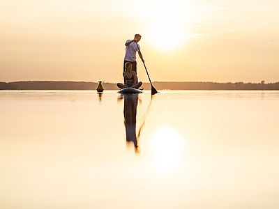Zwei Personen sind auf einem Stand-up-Paddleboard auf einem ruhigen See unterwegs. Eine Person steht und paddelt, die andere sitzt entspannt auf dem Board. Die tief stehende Sonne spiegelt sich auf der glatten Wasseroberfläche und taucht die Szene in warmes, goldenes Licht.