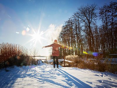 Eine Person in roter Winterjacke steht mit ausgebreiteten Armen auf einer schneebedeckten Wiese. Die tief stehende Sonne scheint durch die kahlen Bäume am Waldrand.