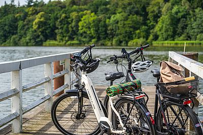 Zwei Fahrräder mit Helmen und Gepäck stehen auf einem Holzsteg am Wasser. Am gegenüberliegenden Ufer erstreckt sich ein dicht bewaldeter, grüner Uferstreifen.