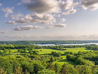 Blick über eine weitläufige Hügellandschaft mit Wiesen, Baumgruppen und Wäldern. Im Hintergrund erstreckt sich ein See entlang des Horizonts. Darüber sind große, teils dichte Wolken am Himmel zu sehen.