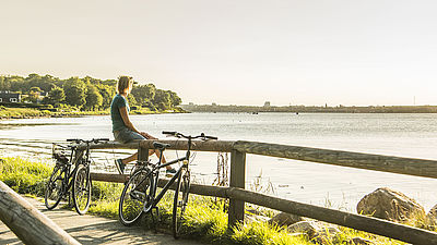 Eine Radfahrerin sitzt auf einem Holzgeländer und schaut auf das Meer. Am Geländer lehnen zwei Fahrräder.