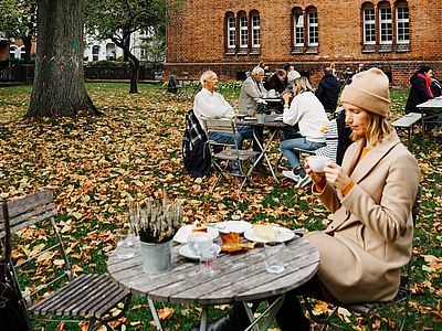 Eine Frau sitzt mit einer Tasse an einem Tisch in einem Café-Garten. Herbstlaub bedeckt den Boden, im Hintergrund sitzen weitere Gäste an Tischen vor einem Backsteingebäude.