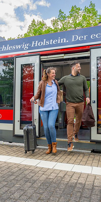Zwei Personen steigen mit Gepäck aus einem roten Regionalzug an einem Bahnhof aus. Die Frau zieht einen Rollkoffer hinter sich her, der Mann trägt eine Reisetasche. Über den Türen des Zuges steht der Schriftzug „Schleswig-Holstein. Der echte Norden.“.