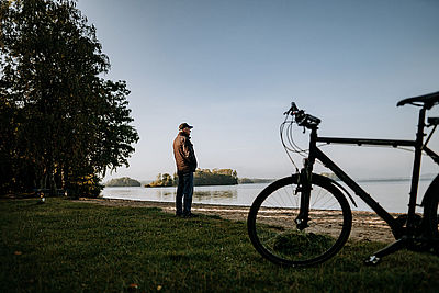 Ein Mann steht an einem kleinen Sandabschnitt und schaut auf einen See. Neben ihm steht ein Fahrrad.