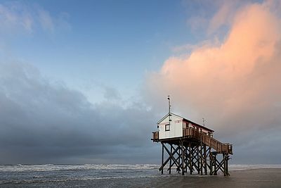 Ein Pfahlbau steht im flachen Wasser vor der Küste. Über dem bewegten Meer treffen dunkle Wolken auf rosa-orange beleuchtete Wolken am Abendhimmel.