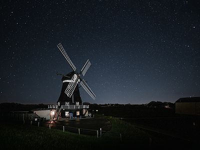 Historische Windmühle mit weißen Flügeln unter einem sternenklaren Nachthimmel. Einzelne Fenster und der Eingangsbereich der Mühle sind beleuchtet.