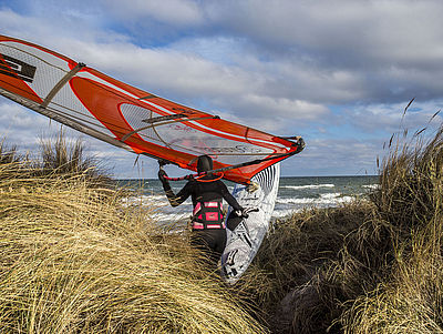 Ein Windsurfer geht mit seinem Board und Segel durch die Dünen in Richtung Ostsee.