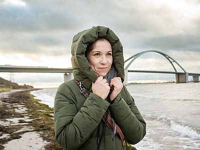 Eine Frau in grüner Winterjacke hält ihre Kapuze mit beiden Händen fest. Im Hintergrund überspannt die Fehmarnsundbrücke das Meer unter einem bewölkten Himmel.