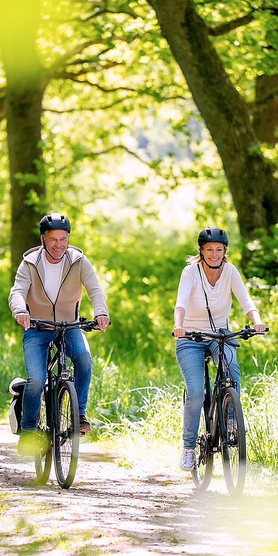Zwei Personen mit Fahrradhelmen fahren auf Fahrrädern einen sonnigen Waldweg entlang. Hohe Bäume und dichtes Grün säumen den Weg.