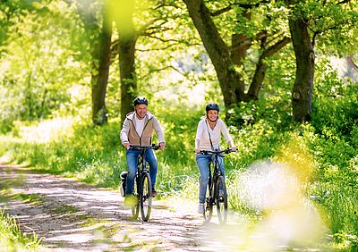 Zwei Personen mit Fahrradhelmen fahren auf Fahrrädern einen sonnigen Waldweg entlang. Hohe Bäume und dichtes Grün säumen den Weg.