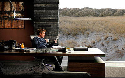 Ein Mann sitzt in einem Büro, trinkt Kaffee und liest Zeitung. Im Hintergrund befinden sich Dünen.